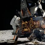 Apollo 12 Achieves Lunar Landing Milestone on This Date On the Moon, an astronaut in a white spacesuit climbs down a ladder on a lunar module. The lunar module is made of metal with gold and silver foil covering some of its lower half.