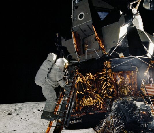 Apollo 12 Achieves Lunar Landing Milestone on This Date On the Moon, an astronaut in a white spacesuit climbs down a ladder on a lunar module. The lunar module is made of metal with gold and silver foil covering some of its lower half.