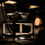 NASA Fine-Tunes MASTER Instrument for Calibration A photo of a room that is mostly dark, with a single light source coming out of a hole in a table. A young engineer with short dark hair looks at the instrument mounted on top of the table, with just his face illuminated by the light.