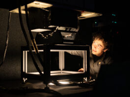NASA Fine-Tunes MASTER Instrument for Calibration A photo of a room that is mostly dark, with a single light source coming out of a hole in a table. A young engineer with short dark hair looks at the instrument mounted on top of the table, with just his face illuminated by the light.