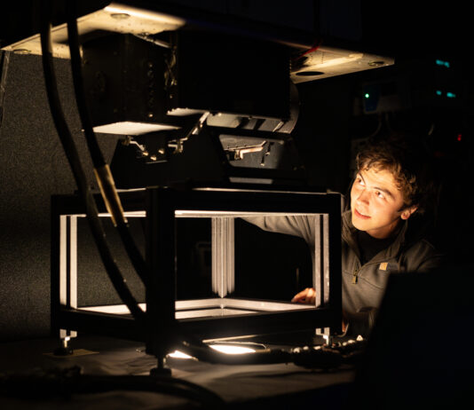 NASA Fine-Tunes MASTER Instrument for Calibration A photo of a room that is mostly dark, with a single light source coming out of a hole in a table. A young engineer with short dark hair looks at the instrument mounted on top of the table, with just his face illuminated by the light.