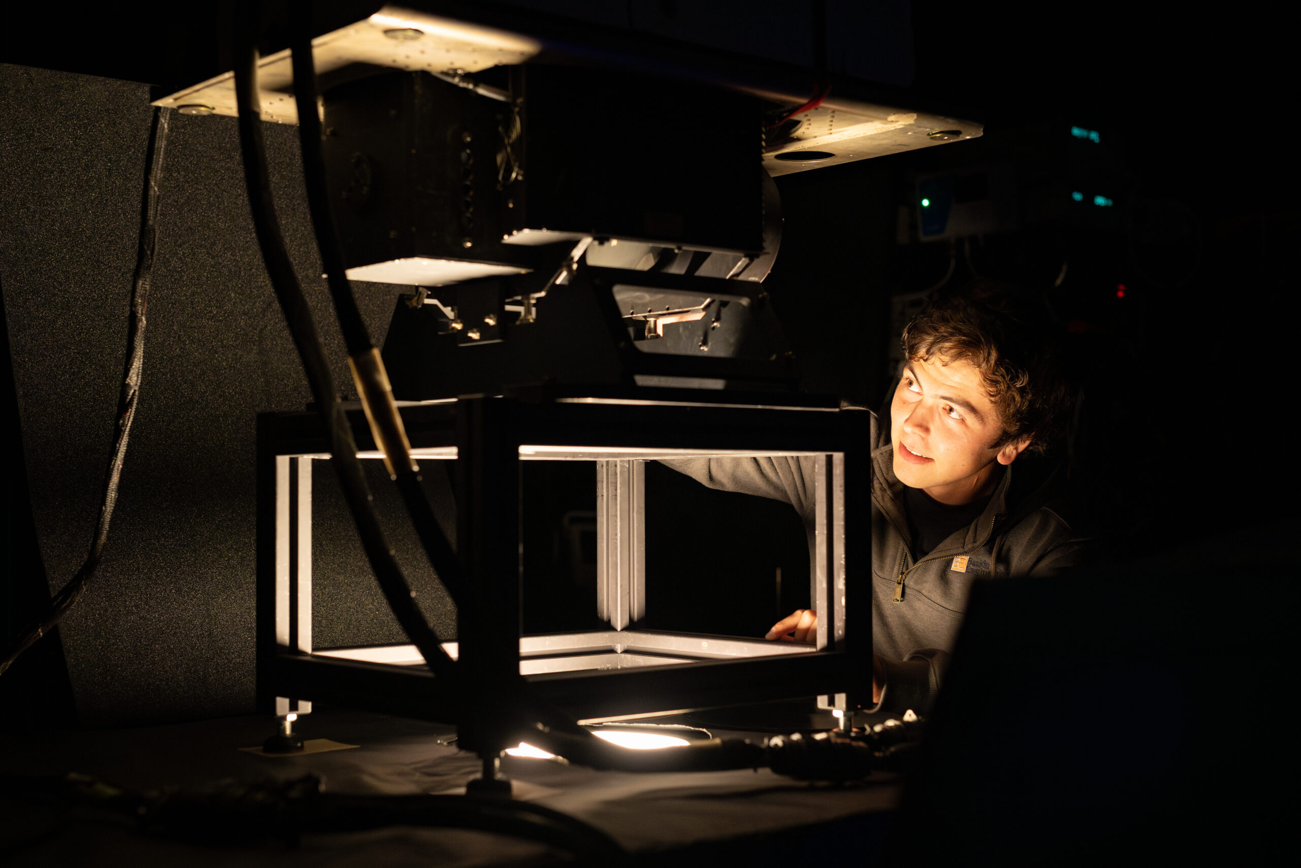 A photo of a room that is mostly dark, with a single light source coming out of a hole in a table. A young engineer with short dark hair looks at the instrument mounted on top of the table, with just his face illuminated by the light.