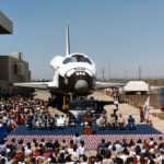Atlantis Space Shuttle Unveiled to Public 40 Years Ago View of a space shuttle orbiter with a about a dozen people on a stage in front of it.