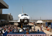 Atlantis Space Shuttle Unveiled to Public 40 Years Ago View of a space shuttle orbiter with a about a dozen people on a stage in front of it.