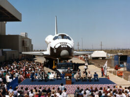 Atlantis Space Shuttle Unveiled to Public 40 Years Ago View of a space shuttle orbiter with a about a dozen people on a stage in front of it.