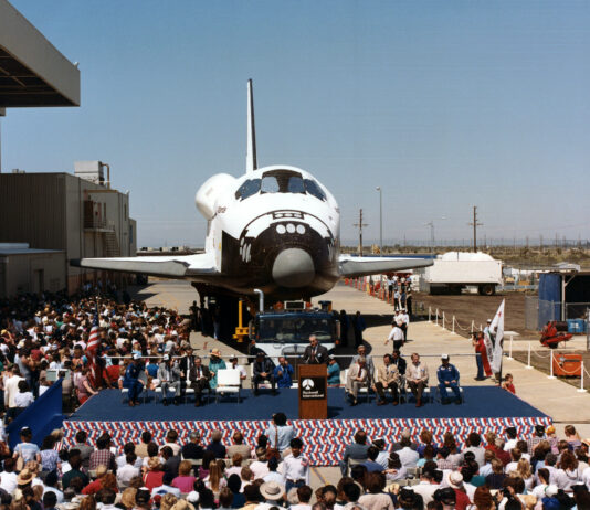 Atlantis Space Shuttle Unveiled to Public 40 Years Ago View of a space shuttle orbiter with a about a dozen people on a stage in front of it.