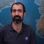 NASA’s Alfonso Delgado Bonal Explores Clouds for Research Alfonso stands in front of a large globe of Earth. He has dark hair and a beard and mustache, with a thoughtful expression. He is wearing a navy blue polo with the blue, red, and white NASA “meatball” logo on the chest patch.