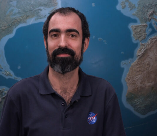 NASA’s Alfonso Delgado Bonal Explores Clouds for Research Alfonso stands in front of a large globe of Earth. He has dark hair and a beard and mustache, with a thoughtful expression. He is wearing a navy blue polo with the blue, red, and white NASA “meatball” logo on the chest patch.