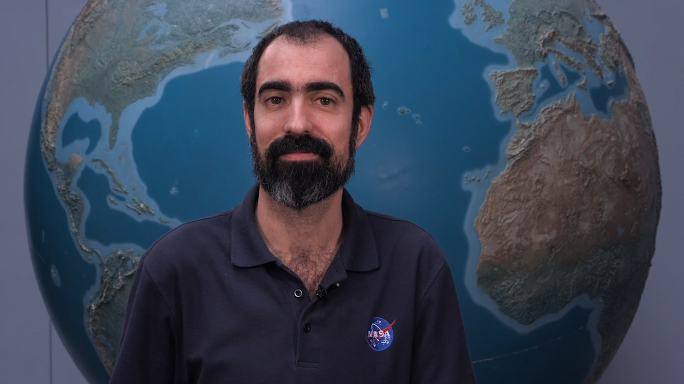bonal2.jpg Alfonso stands in front of a large globe of Earth. He has dark hair and a beard and mustache, with a thoughtful expression. He is wearing a navy blue polo with the blue, red, and white NASA “meatball” logo on the chest patch.