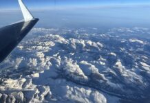 NASA Monitors Snowmelt for Enhanced Water Resource Management A snowy mountain range is seen below through a plane window. The plane’s wing is visible on the left side of the frame, and the sun casts light and shadows across the texture of the landscape. The horizon in the distance is hazy under a blue sky.