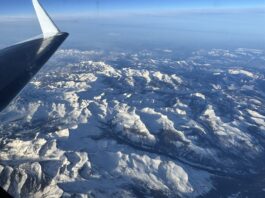 NASA Monitors Snowmelt for Enhanced Water Resource Management A snowy mountain range is seen below through a plane window. The plane’s wing is visible on the left side of the frame, and the sun casts light and shadows across the texture of the landscape. The horizon in the distance is hazy under a blue sky.