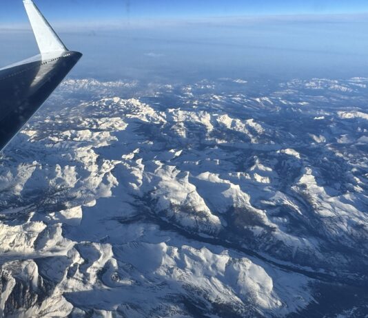 NASA Monitors Snowmelt for Enhanced Water Resource Management A snowy mountain range is seen below through a plane window. The plane’s wing is visible on the left side of the frame, and the sun casts light and shadows across the texture of the landscape. The horizon in the distance is hazy under a blue sky.