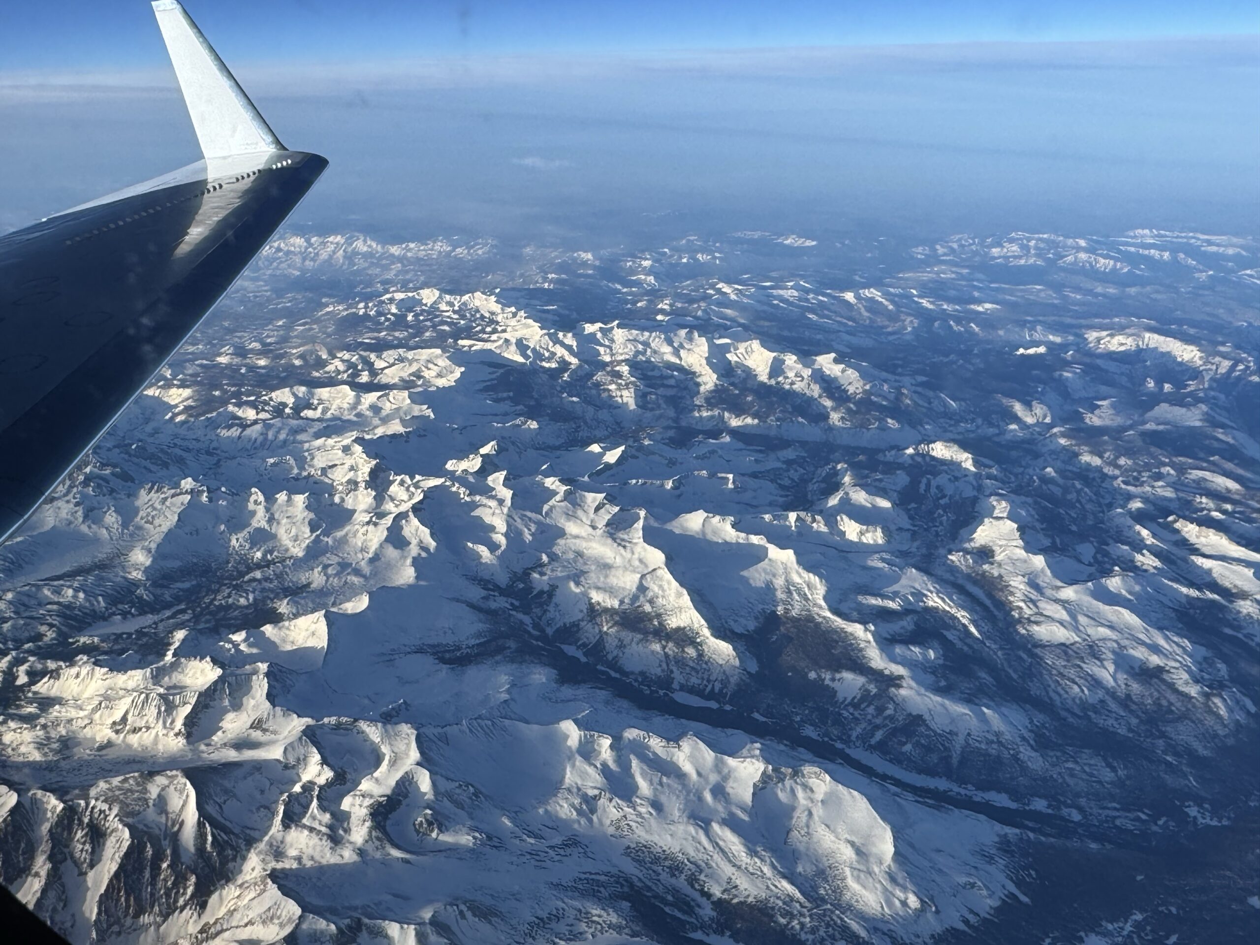 c20a snowstormdust 03172025 05.jpeg A snowy mountain range is seen below through a plane window. The plane’s wing is visible on the left side of the frame, and the sun casts light and shadows across the texture of the landscape. The horizon in the distance is hazy under a blue sky.
