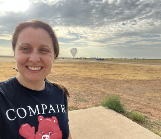 Regina Caputo Shapes the Future of High-Energy Astrophysics at NASA Regina Caputo smiles at the camera in a selfie that captures her head and shoulders. Her brown hair is tied back in a ponytail, and she is wearing a navy-blue tee-shirt which reads “COMPAIR.” Behind Regina is a large open field covered in patches of light brown and yellow grass. The sky is a hazy gray-blue and is covered in dark gray clouds that are thick in certain places but patchy. In the far distance behind Regina, nearing the horizon, is a large space-balloon. The balloon resembles a gray upside-down teardrop, the tip of which just touches the ground.