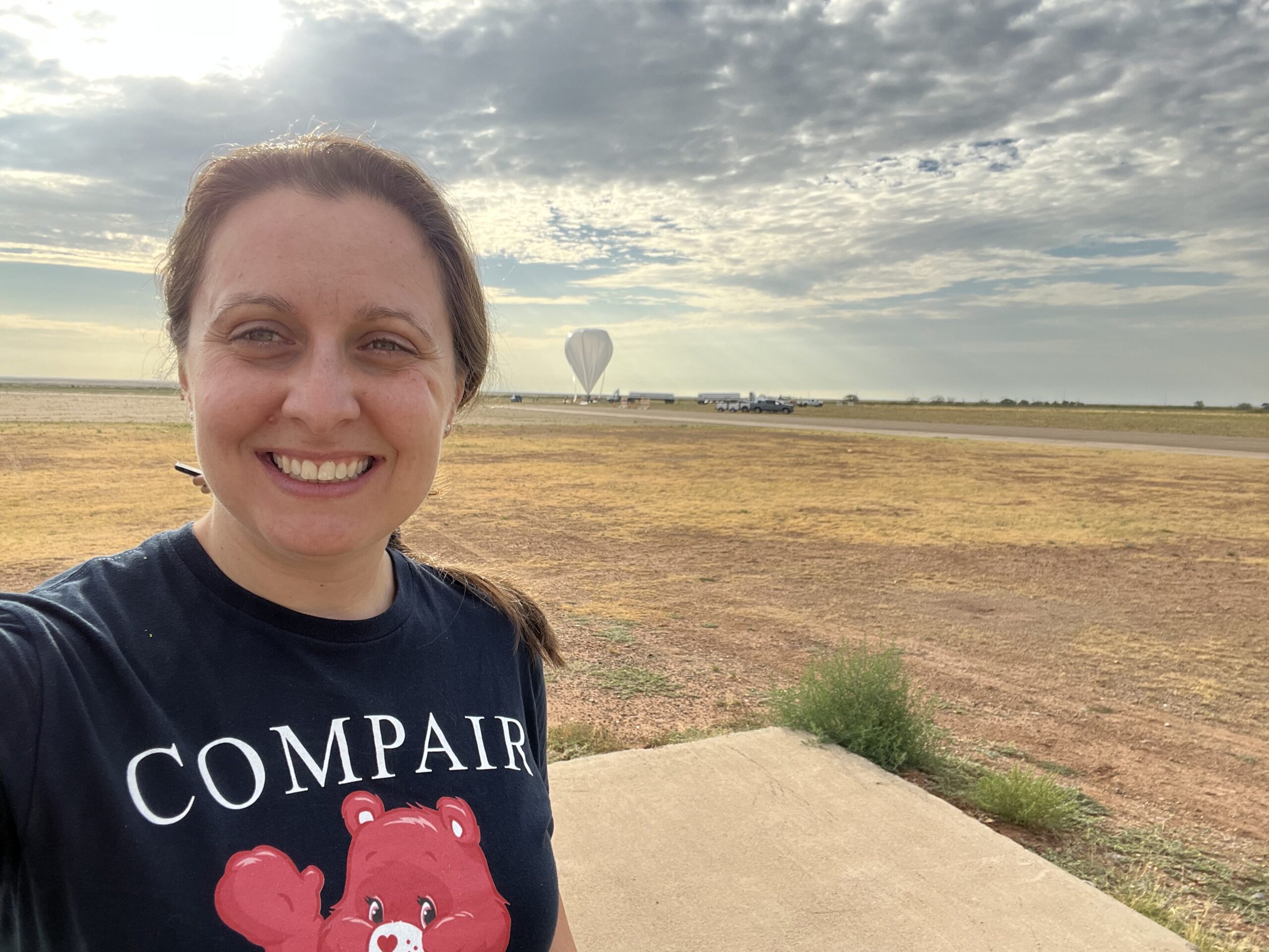 caputo balloon rotated.jpeg Regina Caputo smiles at the camera in a selfie that captures her head and shoulders. Her brown hair is tied back in a ponytail, and she is wearing a navy-blue tee-shirt which reads “COMPAIR.” Behind Regina is a large open field covered in patches of light brown and yellow grass. The sky is a hazy gray-blue and is covered in dark gray clouds that are thick in certain places but patchy. In the far distance behind Regina, nearing the horizon, is a large space-balloon. The balloon resembles a gray upside-down teardrop, the tip of which just touches the ground.