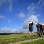 NASA Student Launch Celebrates 25th Anniversary Four people carry a small, thin white rocket across a grassy field. The photo is taken from a lower angle, so the blue sky stretches out over most of the background.