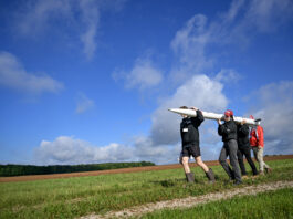 NASA Student Launch Celebrates 25th Anniversary Four people carry a small, thin white rocket across a grassy field. The photo is taken from a lower angle, so the blue sky stretches out over most of the background.