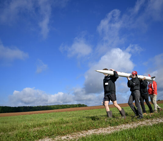 NASA Student Launch Celebrates 25th Anniversary Four people carry a small, thin white rocket across a grassy field. The photo is taken from a lower angle, so the blue sky stretches out over most of the background.