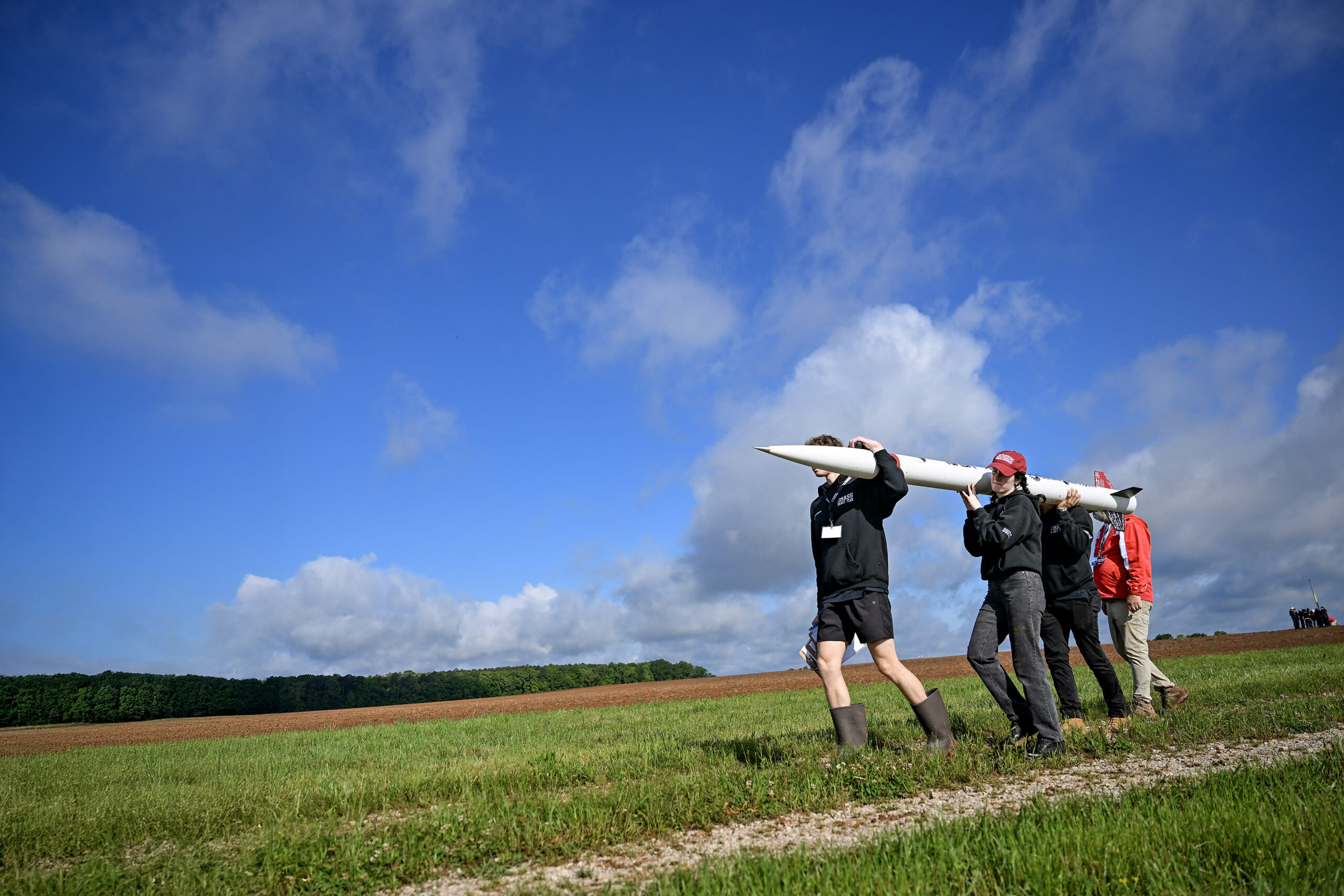 ceb 0969orig.jpg Four people carry a small, thin white rocket across a grassy field. The photo is taken from a lower angle, so the blue sky stretches out over most of the background.