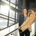 NASA’s Tami Wisniewski: Manager and Program Analyst Tami, a woman of Native American heritage from the Delaware Nation, smiles standing on a staircase in a modern, sunlit building, holding a traditional handcrafted cradleboard adorned with intricate beadwork. She is wearing a paisley patterned shirt covered with a black sweater that has the NASA insignia on the right breast.