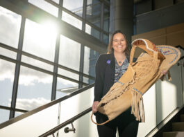 NASA’s Tami Wisniewski: Manager and Program Analyst Tami, a woman of Native American heritage from the Delaware Nation, smiles standing on a staircase in a modern, sunlit building, holding a traditional handcrafted cradleboard adorned with intricate beadwork. She is wearing a paisley patterned shirt covered with a black sweater that has the NASA insignia on the right breast.