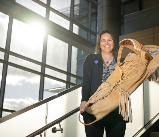 NASA’s Tami Wisniewski: Manager and Program Analyst Tami, a woman of Native American heritage from the Delaware Nation, smiles standing on a staircase in a modern, sunlit building, holding a traditional handcrafted cradleboard adorned with intricate beadwork. She is wearing a paisley patterned shirt covered with a black sweater that has the NASA insignia on the right breast.
