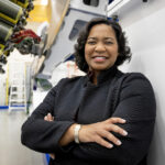 NASA’s Tawnya Plummer Laughinghouse Leads Laboratory Tawnya Laughinghouse is standing in a high-tech industrial facility, smiling and crossing her arms. She is wearing a textured black jacket and a gold bracelet. Behind her are large pieces of machinery and industrial equipment, with bright lighting highlighting the space.