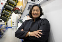 NASA’s Tawnya Plummer Laughinghouse Leads Laboratory Tawnya Laughinghouse is standing in a high-tech industrial facility, smiling and crossing her arms. She is wearing a textured black jacket and a gold bracelet. Behind her are large pieces of machinery and industrial equipment, with bright lighting highlighting the space.