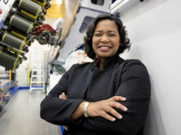 NASA’s Tawnya Plummer Laughinghouse Leads Laboratory Tawnya Laughinghouse is standing in a high-tech industrial facility, smiling and crossing her arms. She is wearing a textured black jacket and a gold bracelet. Behind her are large pieces of machinery and industrial equipment, with bright lighting highlighting the space.