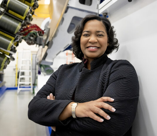 NASA’s Tawnya Plummer Laughinghouse Leads Laboratory Tawnya Laughinghouse is standing in a high-tech industrial facility, smiling and crossing her arms. She is wearing a textured black jacket and a gold bracelet. Behind her are large pieces of machinery and industrial equipment, with bright lighting highlighting the space.