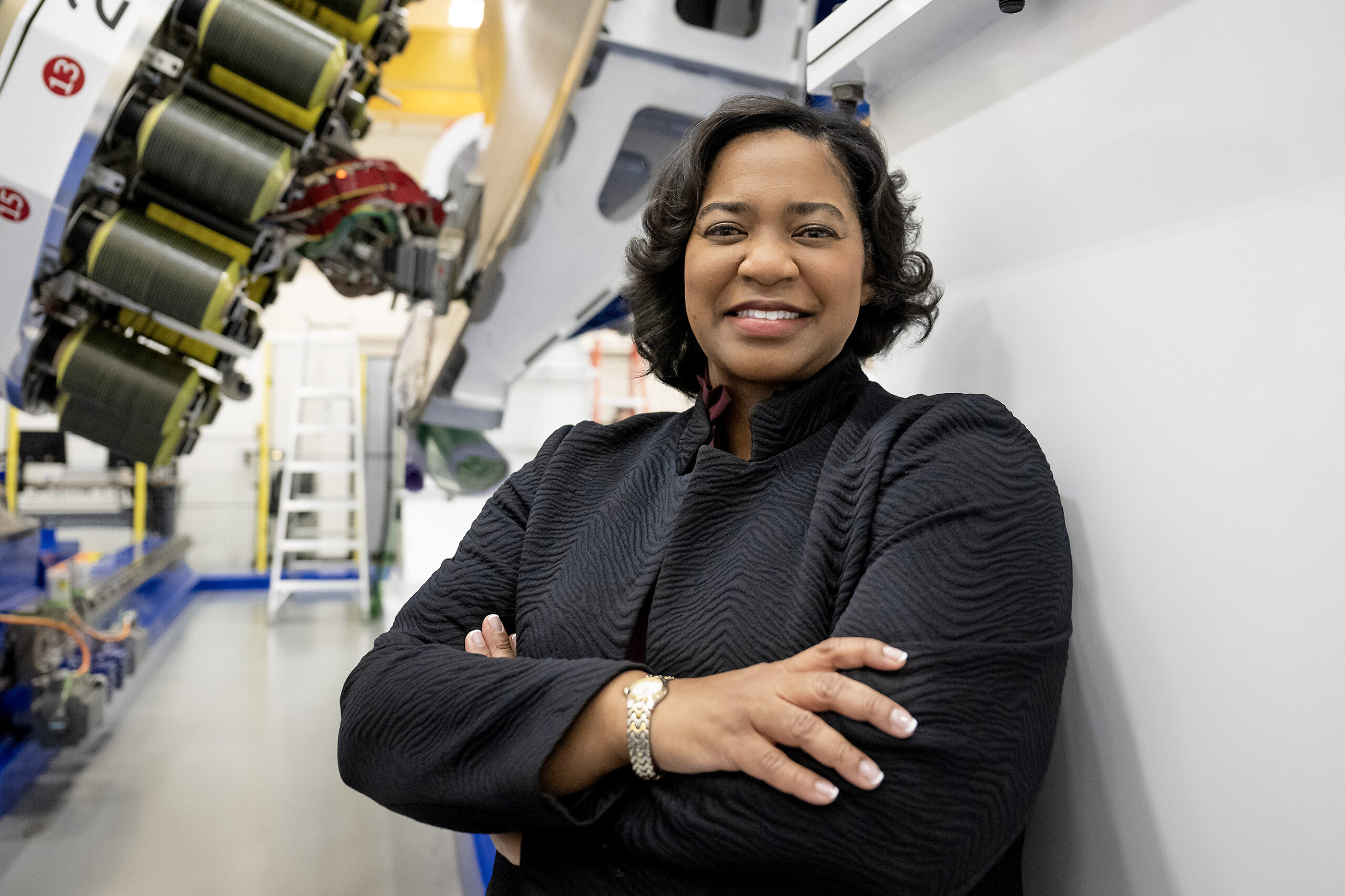 ceb 5997a.jpg Tawnya Laughinghouse is standing in a high-tech industrial facility, smiling and crossing her arms. She is wearing a textured black jacket and a gold bracelet. Behind her are large pieces of machinery and industrial equipment, with bright lighting highlighting the space.