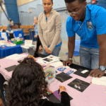NASA eClips Ambassadors Illuminate CNU’s 2025 STEM Day Event Two STEM Student Ambassadors, one a female wearing a tan jacket and one a male with glasses wearing his turquoise Ambassador polo shirt look over an exhibit table laden with black construction paper and pastels and explain to a young female with curly dark hair and a black shirt with pink bracelet as she draws her own aurora using a purple stencil on a piece of black construction paper.