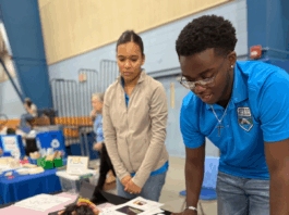 NASA eClips Ambassadors Illuminate CNU’s 2025 STEM Day Event Two STEM Student Ambassadors, one a female wearing a tan jacket and one a male with glasses wearing his turquoise Ambassador polo shirt look over an exhibit table laden with black construction paper and pastels and explain to a young female with curly dark hair and a black shirt with pink bracelet as she draws her own aurora using a purple stencil on a piece of black construction paper.