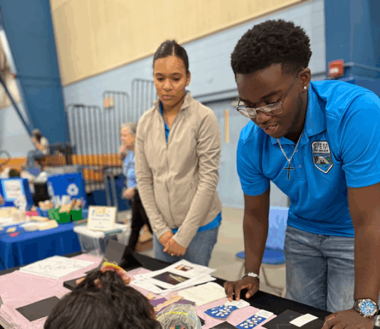 NASA eClips Ambassadors Illuminate CNU’s 2025 STEM Day Event Two STEM Student Ambassadors, one a female wearing a tan jacket and one a male with glasses wearing his turquoise Ambassador polo shirt look over an exhibit table laden with black construction paper and pastels and explain to a young female with curly dark hair and a black shirt with pink bracelet as she draws her own aurora using a purple stencil on a piece of black construction paper.