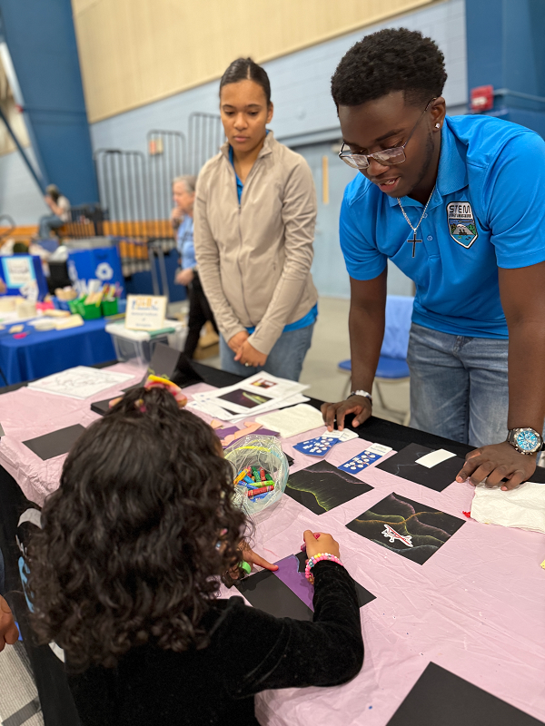 Two STEM Student Ambassadors, one a female wearing a tan jacket and one a male with glasses wearing his turquoise Ambassador polo shirt look over an exhibit table laden with black construction paper and pastels and explain to a young female with curly dark hair and a black shirt with pink bracelet as she draws her own aurora using a purple stencil on a piece of black construction paper.