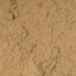 Curiosity Rover Finds Mars’ Largest Organic Molecules Yet This animated image shows a close-up view of a flat, dusty, tan-colored rock surface with fine cracks and small, dark specks scattered across it. In the animation, a hole appears in the center of the frame, with loose powdered material surrounding the hole. The surrounding rock surface remains unchanged, highlighting the freshly drilled hole and the fine texture of the rock.