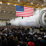 NASA Forms Partnership with Private Sector to Boost Mars Research NASA Administrator Jared Isaacman stands in front of a large, cylindrical, gray spacecraft with a white Relativity logo on it and addresses hundreds of Relativity Space employees. A large American flag hangs from the ceiling.