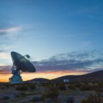 Introducing Lindsai Bland from NASA’s Space Operations Team A large antenna, part of the Deep Space Network, sits on top of a hill at sunset. The sky around the antenna is a bright orange but bleeds into a dark blue around the edges of the image.