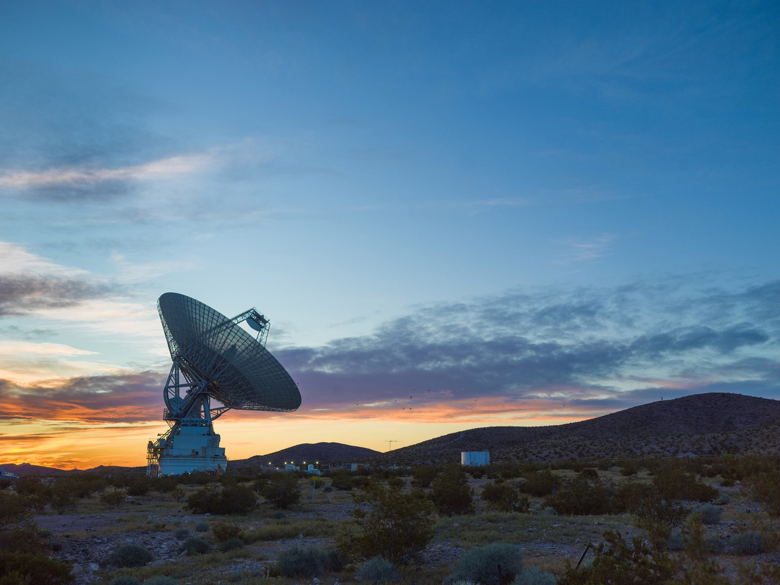 A large antenna, part of the Deep Space Network, sits on top of a hill at sunset. The sky around the antenna is a bright orange but bleeds into a dark blue around the edges of the image.