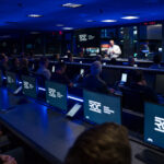 NASA JPL Launches New Center for Lunar and Martian Missions A man, illuminated by white light, talking to a group of people in a room that is otherwise dark and dimly lit with blue light.