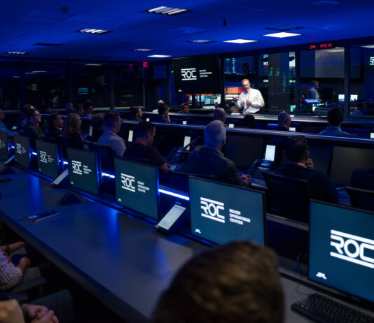 NASA JPL Launches New Center for Lunar and Martian Missions A man, illuminated by white light, talking to a group of people in a room that is otherwise dark and dimly lit with blue light.