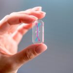 NASA’s Artemis II to Test Avatars for Astronaut Health Two fingers holding a transparent rectangular device the size of a USB drive. Inside the device is a long line of blue with two short, red lines at opposite ends.