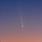 Rare Guest Visits NASA’s Michoud Facility The night sky goes from blue to pink as you look from top to bottom. In the middle is a comet; it looks like a relatively bright yellow dot with a long, diffuse yellow tail. Some stars are visible in the blue portion of the sky.