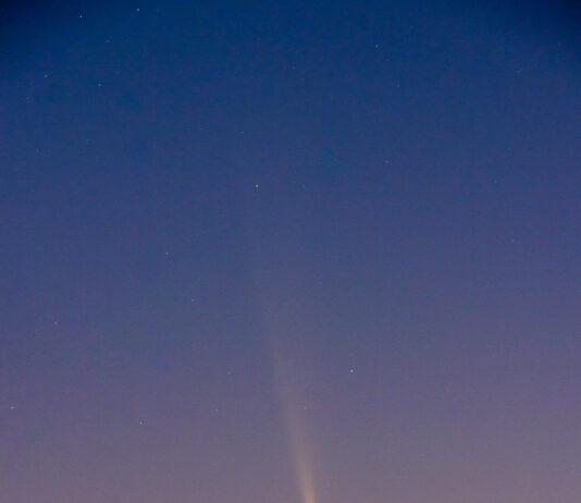 Rare Guest Visits NASA’s Michoud Facility The night sky goes from blue to pink as you look from top to bottom. In the middle is a comet; it looks like a relatively bright yellow dot with a long, diffuse yellow tail. Some stars are visible in the blue portion of the sky.
