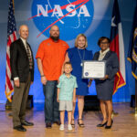Catherine Staggs: Enhancing Artemis with NASA Contracting Skills A blonde woman in a knee-length blue dress receives an award from the director of Johnson Space Center, on stage with her husband and young son.
