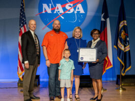Catherine Staggs: Enhancing Artemis with NASA Contracting Skills A blonde woman in a knee-length blue dress receives an award from the director of Johnson Space Center, on stage with her husband and young son.