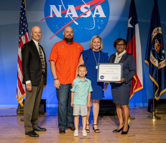 Catherine Staggs: Enhancing Artemis with NASA Contracting Skills A blonde woman in a knee-length blue dress receives an award from the director of Johnson Space Center, on stage with her husband and young son.