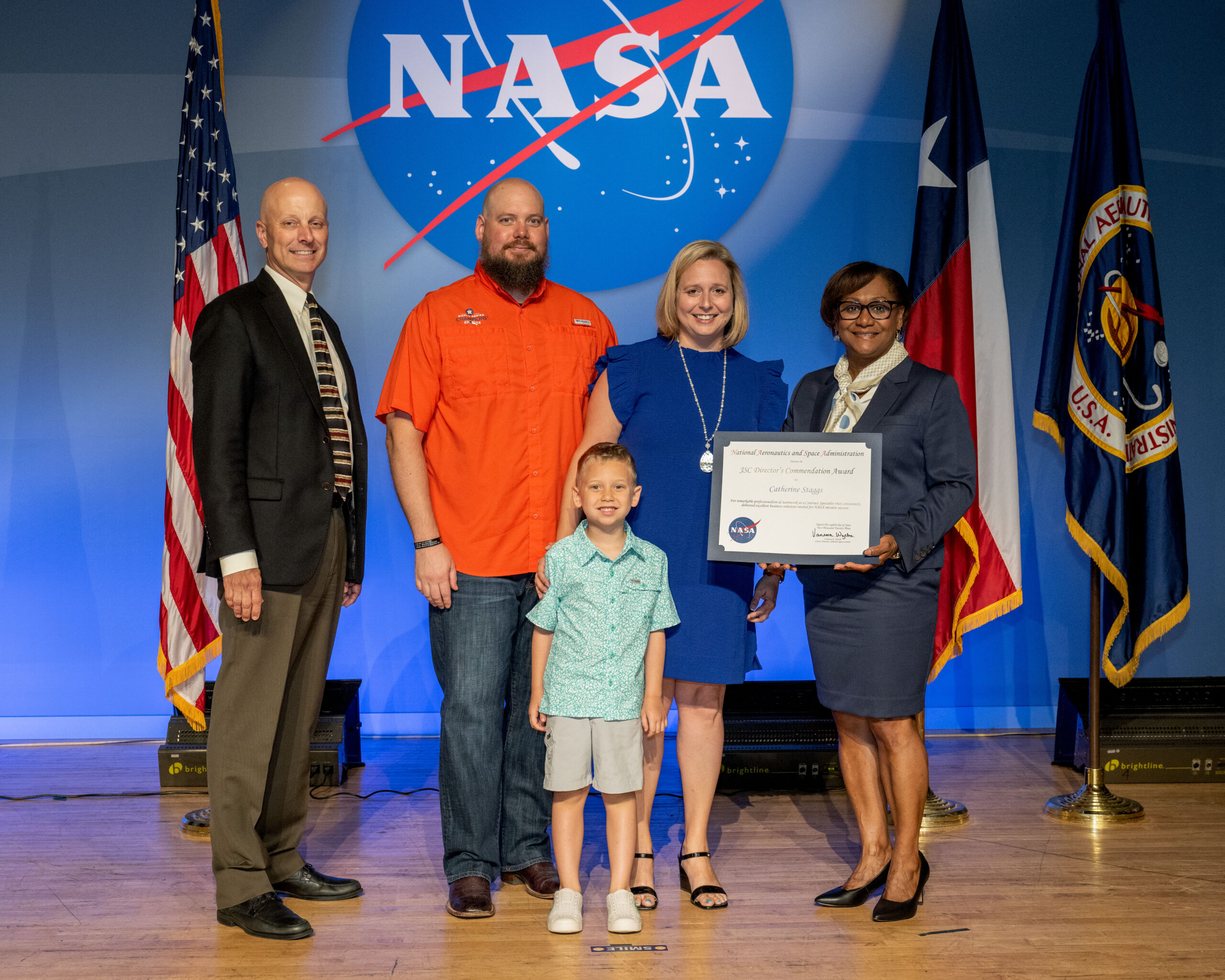 family picture nasa 002.jpg A blonde woman in a knee-length blue dress receives an award from the director of Johnson Space Center, on stage with her husband and young son.