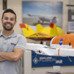 NASA Highlights: Inspiring Engineer Felipe Valdez A man wearing a gray NASA shirt posing to the left of a subscale model or an aircraft that is orange and white with the NASA meatball on it. In the background, there are aviation posters located on the wall and another subscale model on display that is yellow and red.