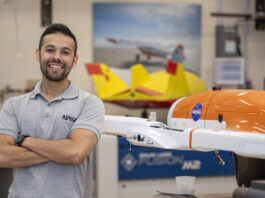 Celebrating Felipe Valdez: An Inspiring NASA Engineer A man wearing a gray NASA shirt posing to the left of a subscale model or an aircraft that is orange and white with the NASA meatball on it. In the background, there are aviation posters located on the wall and another subscale model on display that is yellow and red.